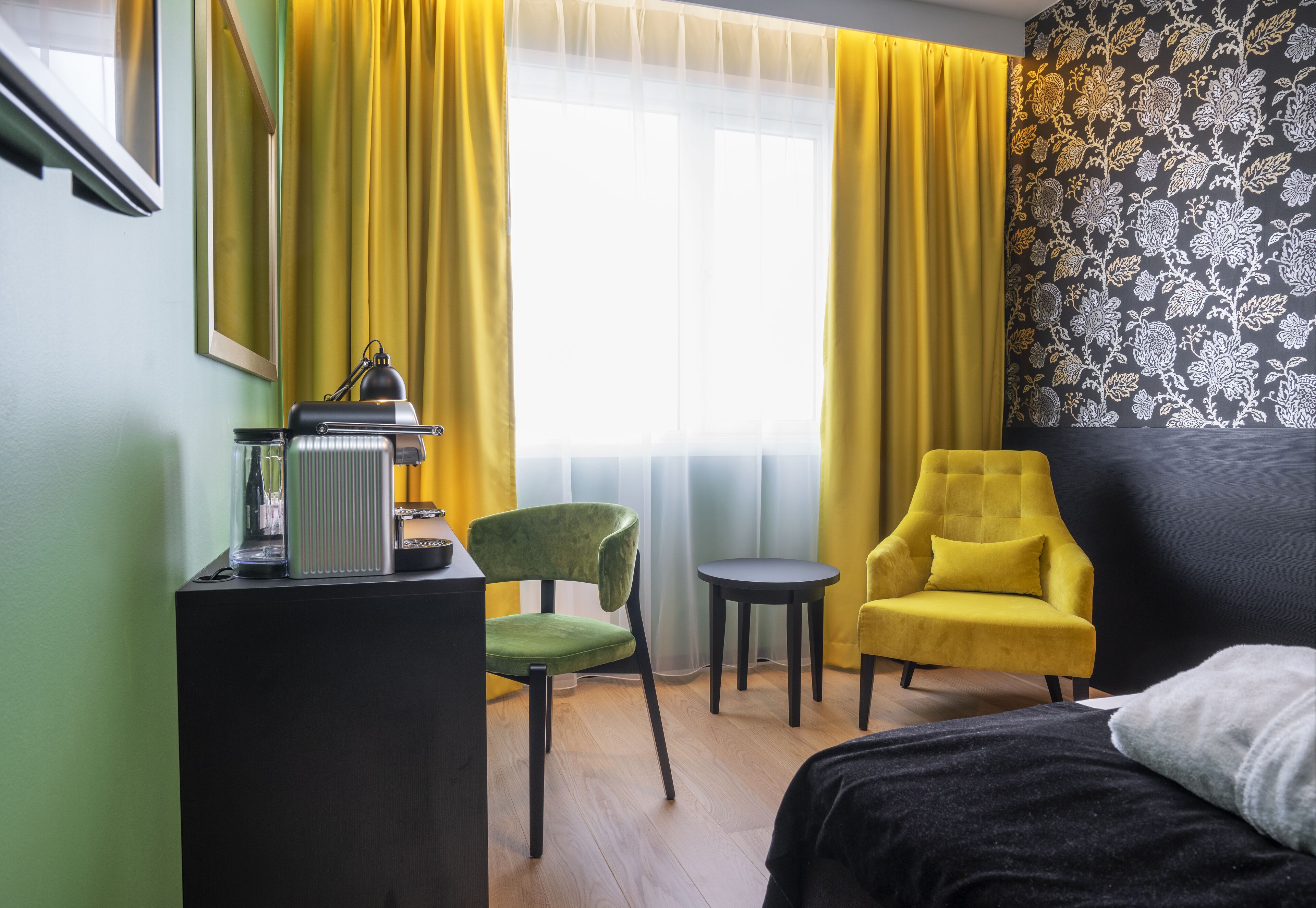 A hotel room with a bed, a desk with a lamp, a green chair, a yellow armchair, and a small table.