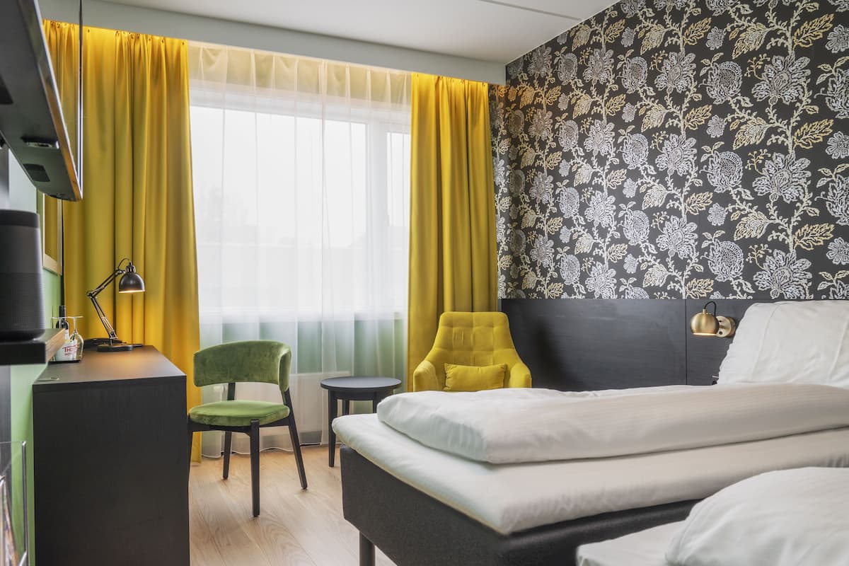 Quarto de hotel com uma cama, uma escrivaninha com luminária, uma cadeira e uma pequena mesa.