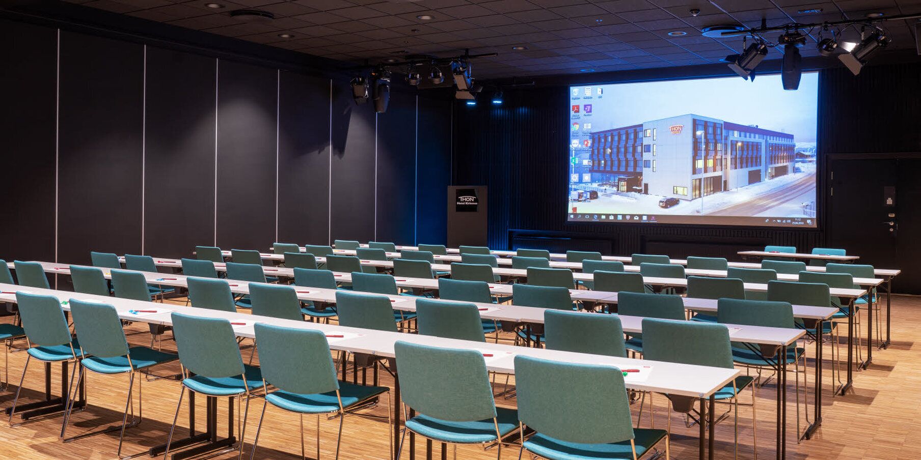 Une salle de conférence avec des rangées de tables et de chaises, un grand écran affichant une image d’un bâtiment, et un pupitre avec une pancarte.