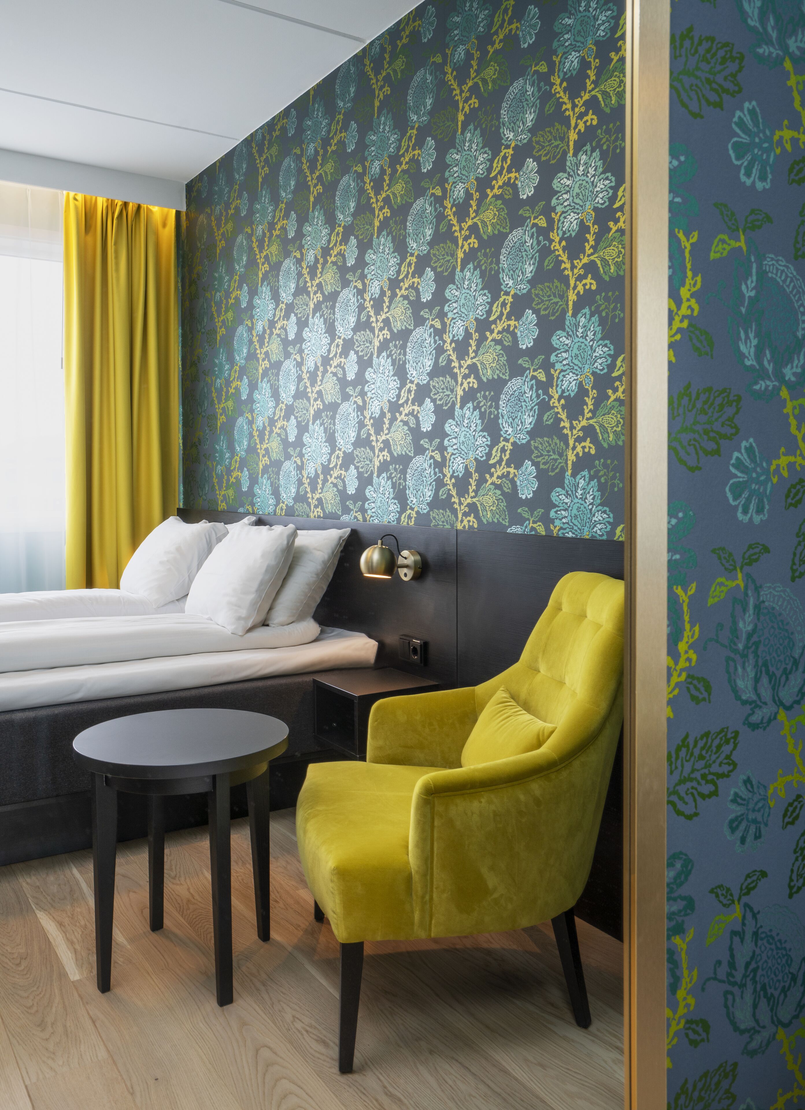 A modern hotel room with a bed, a yellow armchair, a small round table, and a patterned wallpaper.
