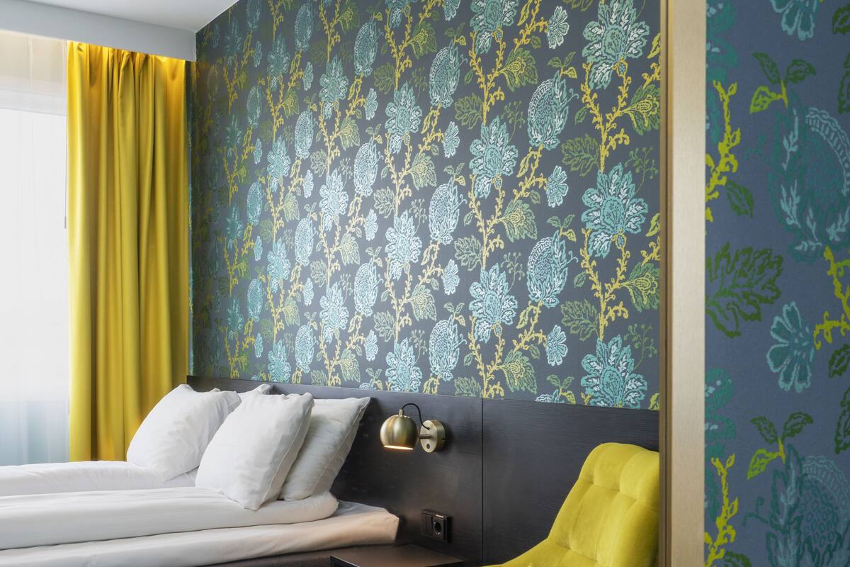 Una habitación de hotel moderna con una cama, un sillón amarillo, una mesita redonda y un papel tapiz estampado.