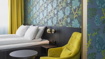 A modern hotel room with a bed, a yellow armchair, a small round table, and a patterned wallpaper.