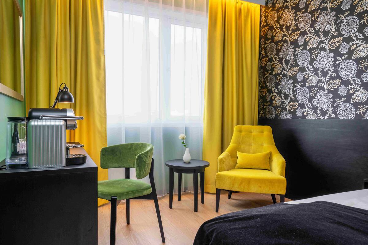 Quarto de hotel com uma cama, uma escrivaninha com luminária, uma cadeira verde, uma poltrona amarela e uma pequena mesa com um vaso.