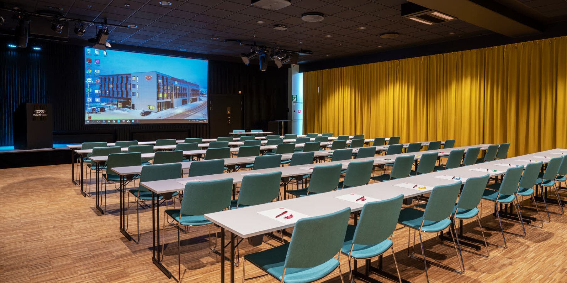 A conference room with rows of tables and chairs, a large screen displaying a building, and a yellow curtain backdrop.