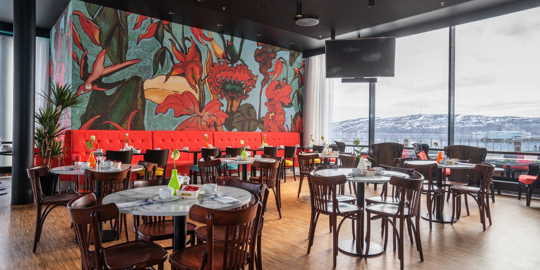 A restaurant with round tables and chairs, a red cushioned bench, and a large mural of flowers on the wall.