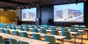 Uma sala de conferências com fileiras de mesas e cadeiras, grandes telas exibindo um edifício e um palco com um púlpito.