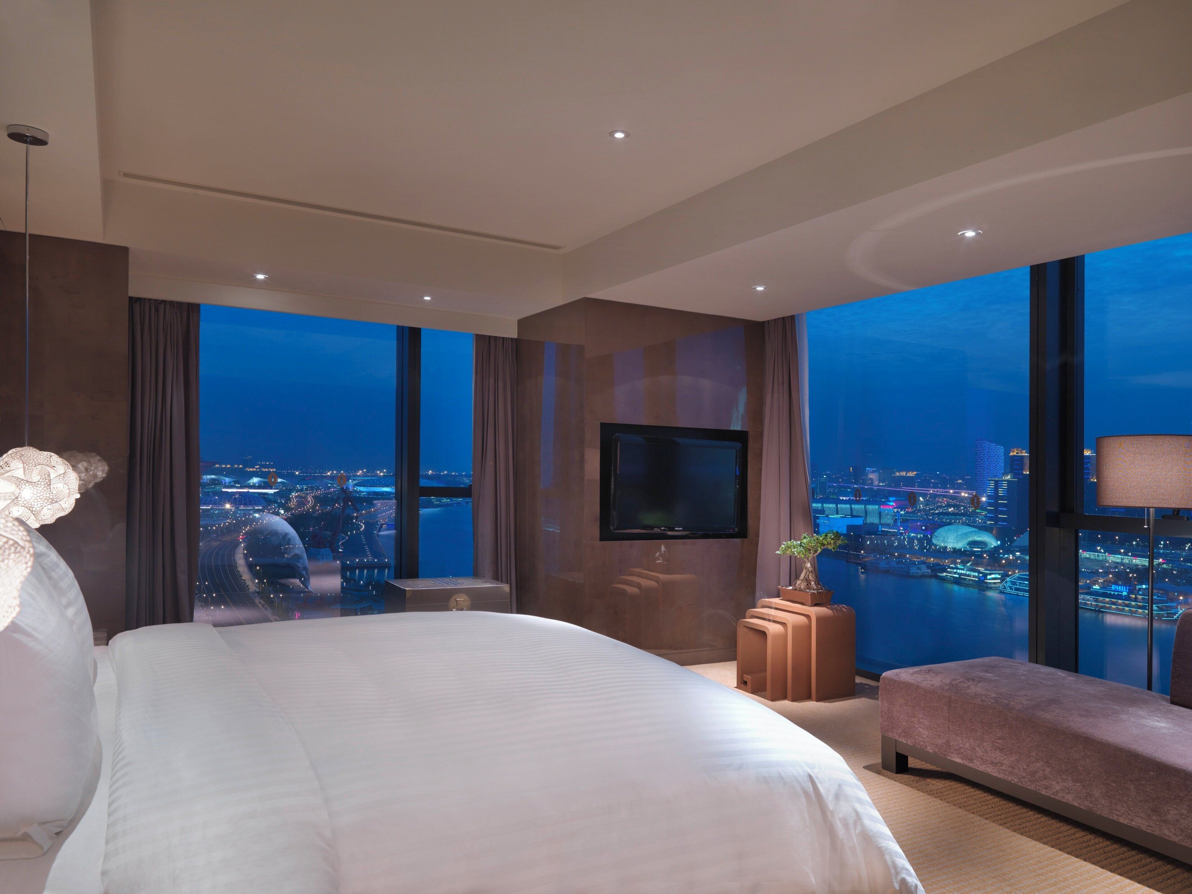 A modern hotel room with a large bed, a flat-screen TV, a seating area, and a view of a cityscape at night.
