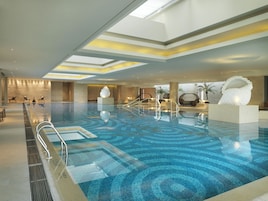 Un'ampia piscina interna dal design moderno, caratterizzata da un motivo geometrico sul pavimento e un'area centrale con posti a sedere e una grande scultura bianca.