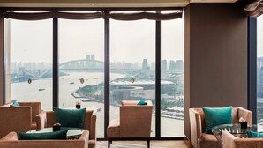 Una moderna camera d'hotel con ampie finestre a tutta altezza che offrono vista sulla città, con un fiume e un ponte. Ci sono due divani con cuscini verdi e un piccolo tavolino con piano in vetro.