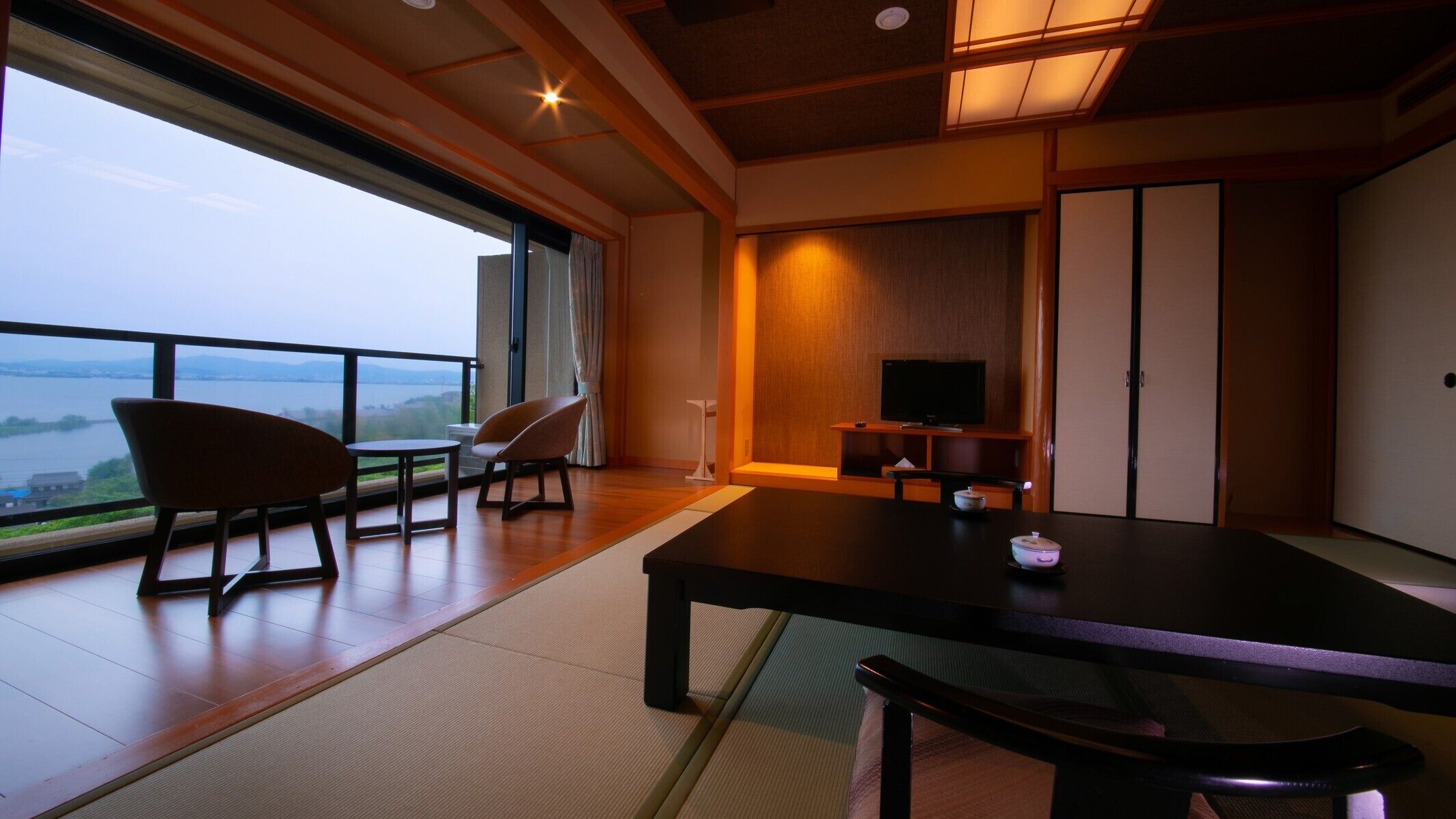 A room with a large window overlooking a body of water, a television on a stand, and a tatami mat floor.