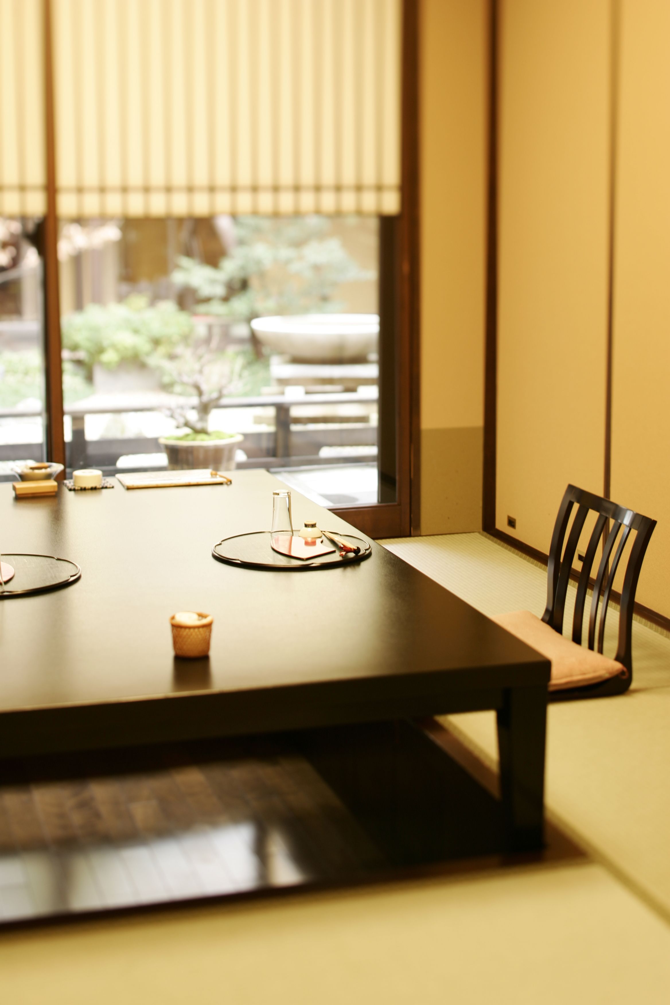 Una mesa de comedor con una silla, una ventana con persianas y vistas al exterior con vegetación.