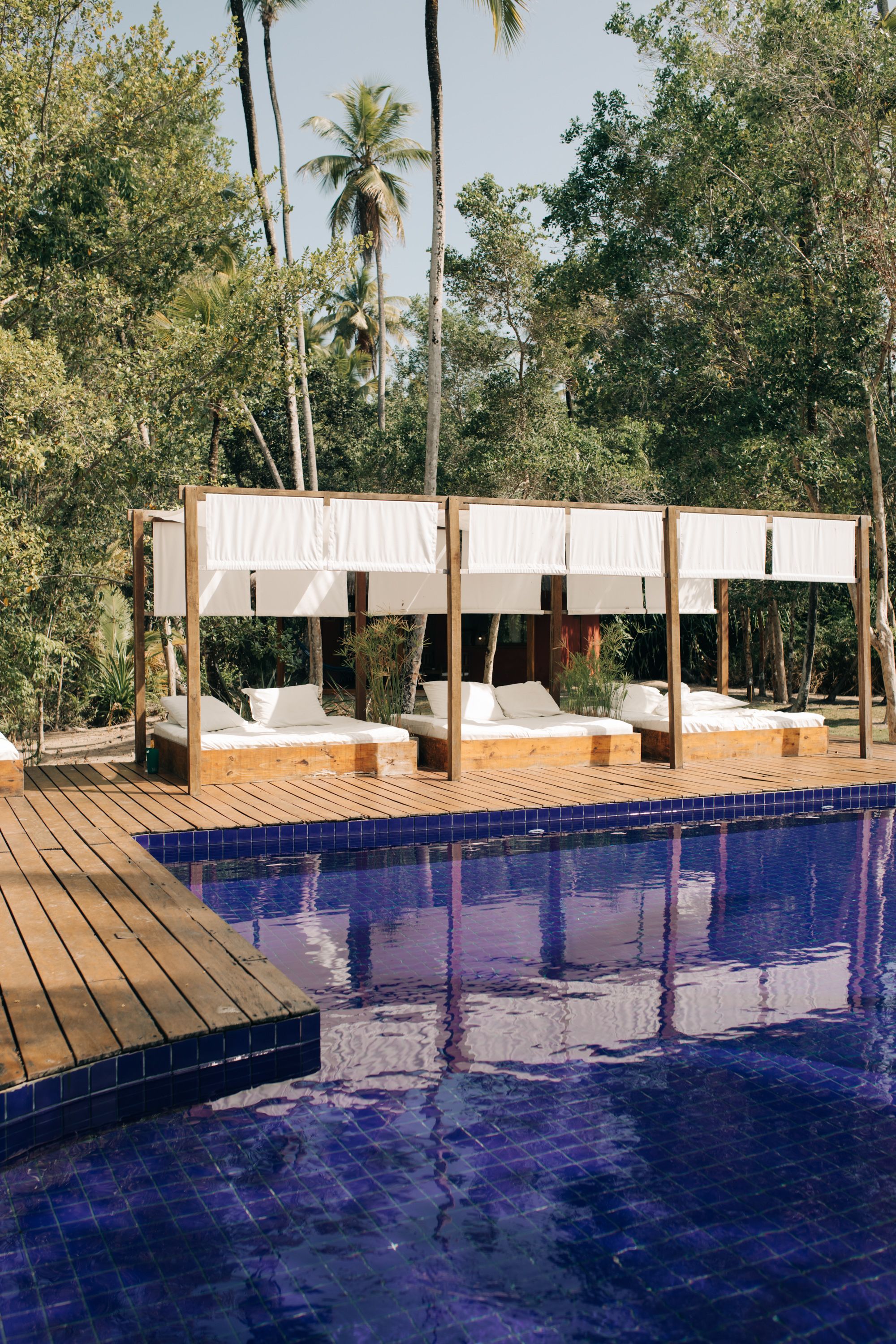 Zona junto a la piscina con terraza de madera, dos camas con dosel blanco y una piscina azul cristalina.