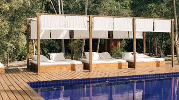 Zona junto a la piscina con terraza de madera, dos camas con dosel blanco y una piscina azul cristalina.