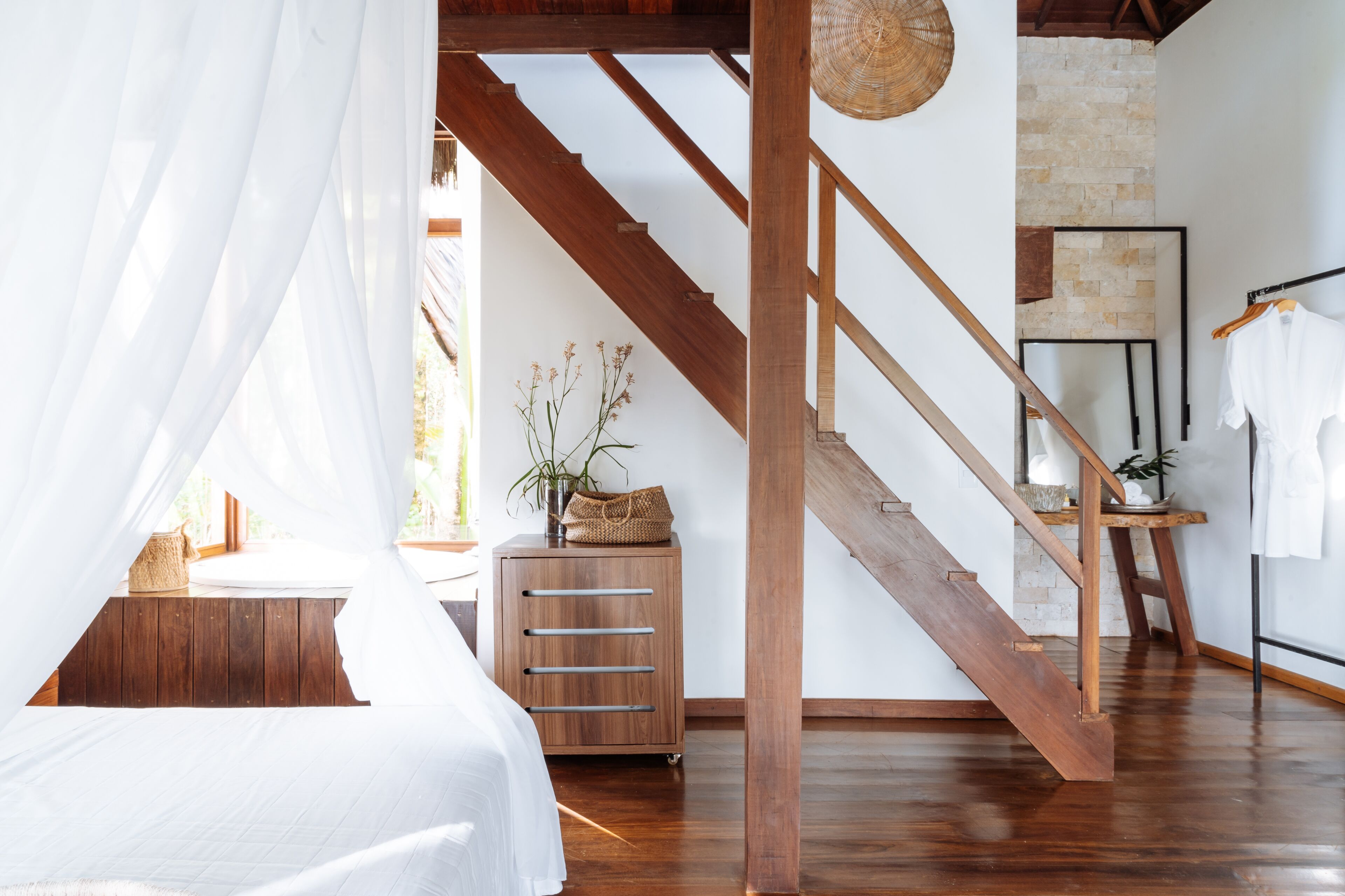 A bedroom with a wooden staircase, a bed with white bedding, and a small wooden cabinet with drawers.