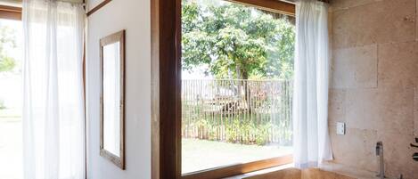 A bathroom with a bathtub, a window with curtains, and a view of a garden.
