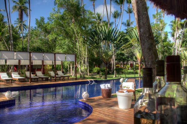 Un área de piscina con sillones de descanso, una terraza de madera y un exuberante jardín tropical de fondo.