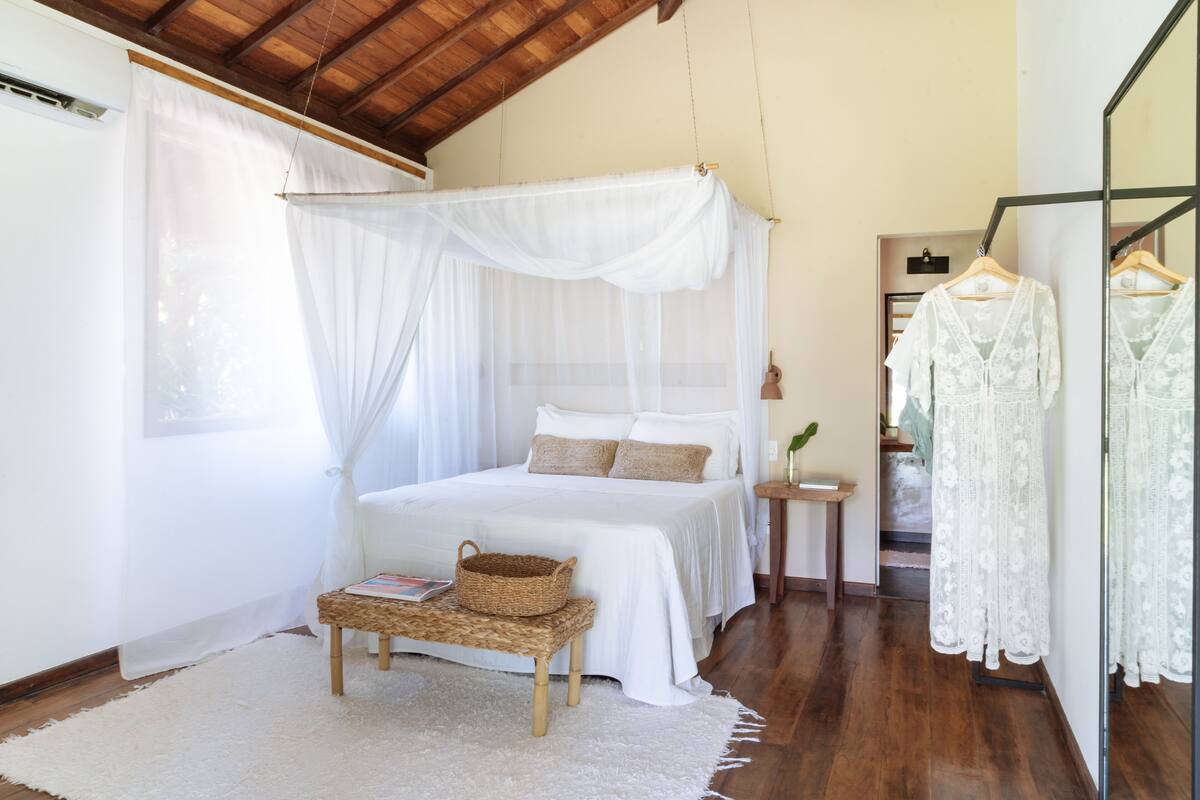 A bedroom with a canopy bed, a bedside table with a lamp, a wicker basket, and a hanging dress rack.