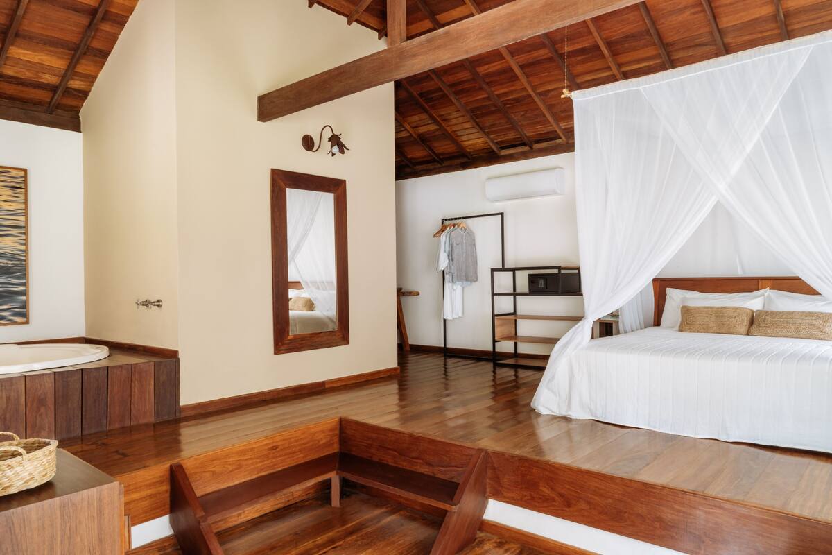 A bedroom with a wooden floor, a bed with white bedding, a bedside table, a mirror, and a hanging light fixture.