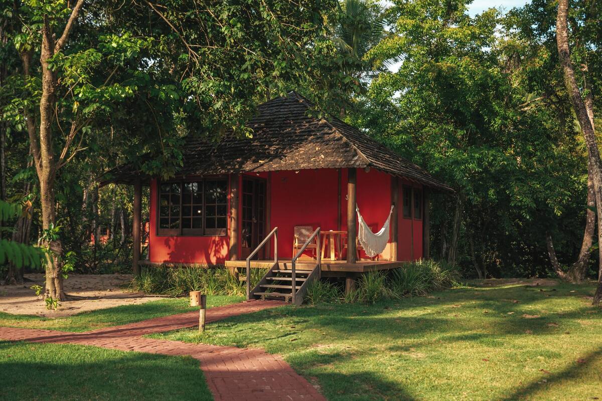 A red wooden cabin with a porch and hammock, surrounded by lush greenery.