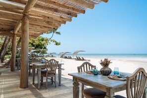 Terraza de madera con mesas y sillas, con vista a una playa con sombrillas y tumbonas.