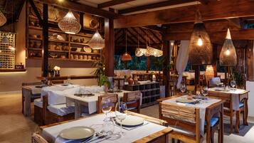 A restaurant with wooden tables and chairs, hanging woven light fixtures, and shelves with pottery and plants.