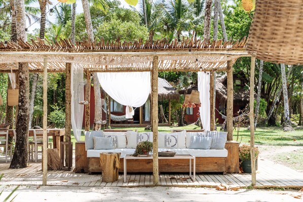 Un área de estar al aire libre con muebles de madera y un toldo, rodeada de abundante vegetación y una estructura con techo de paja al fondo.
