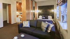 A modern hotel room with a dark-colored sofa, a bedside table with decorative items, a large bed with multiple pillows, a wooden wardrobe, and a kitchenette in the background. - Warsaw Apartments Magnolie (Warsaw)