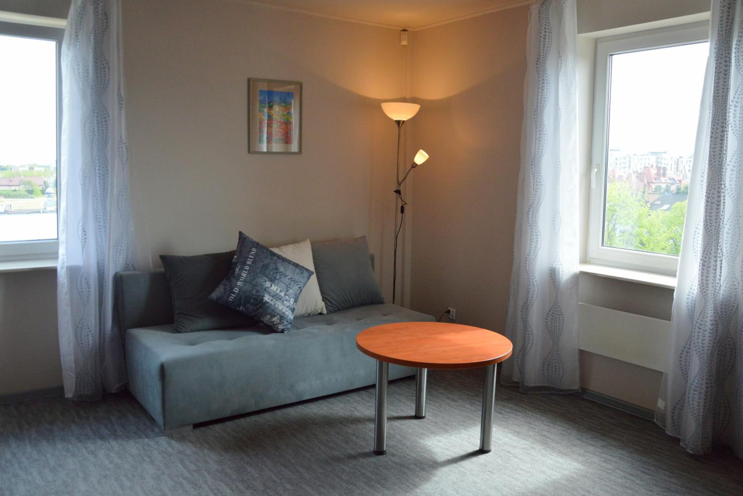 Une pièce avec un canapé gris, une petite table ronde, un lampadaire et une fenêtre avec des rideaux.