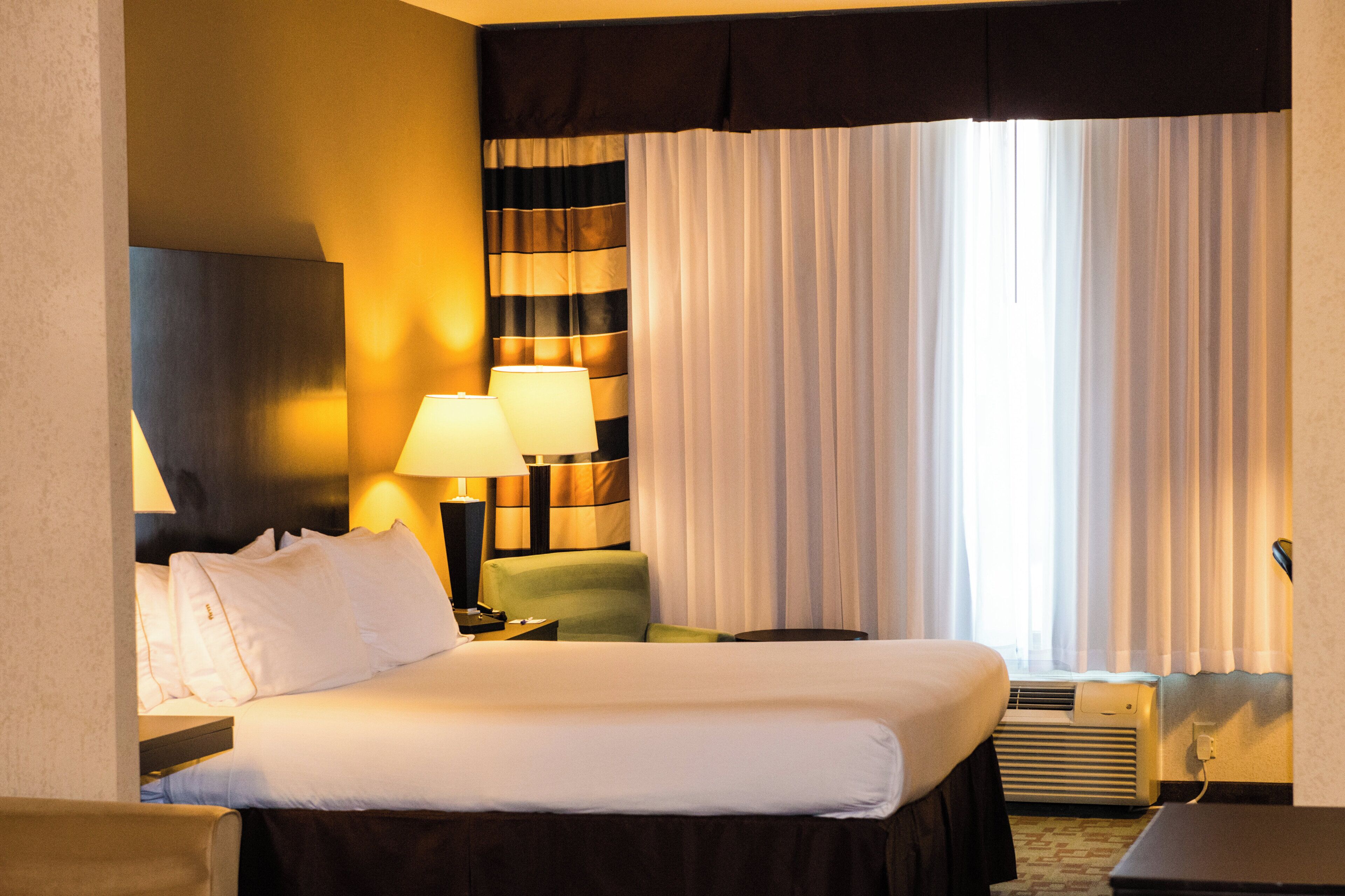 A hotel room with a bed, two bedside lamps, a chair, a nightstand, and a window with curtains.