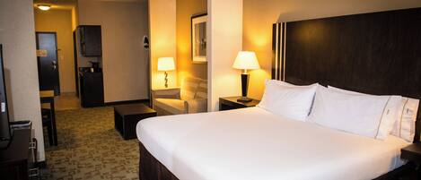 A hotel room with a large bed, a desk, a chair, a TV, and a small kitchenette.