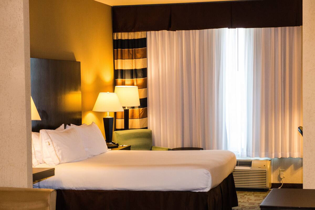 Habitación de hotel con cama, dos lámparas de noche, un sillón verde, aire acondicionado instalado en la pared y una ventana con cortinas.