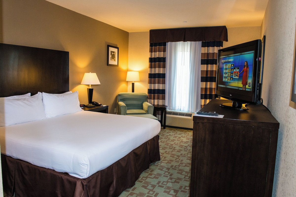 Una habitación de hotel con una cama grande, televisión, una silla, un escritorio y una ventana con cortinas.