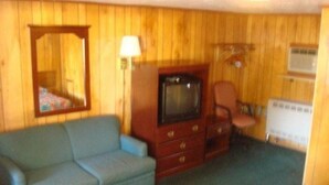 A room with wooden panel walls, a blue sofa, a wooden cabinet with drawers, a television, a chair, and a radiator. - Elmwood Motor Lodge (Boscawen)