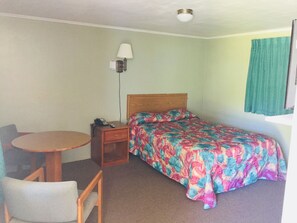 A small, simple room with a bed, a bedside table with a lamp, a chair, and a small table. - Elmwood Motor Lodge (Boscawen)