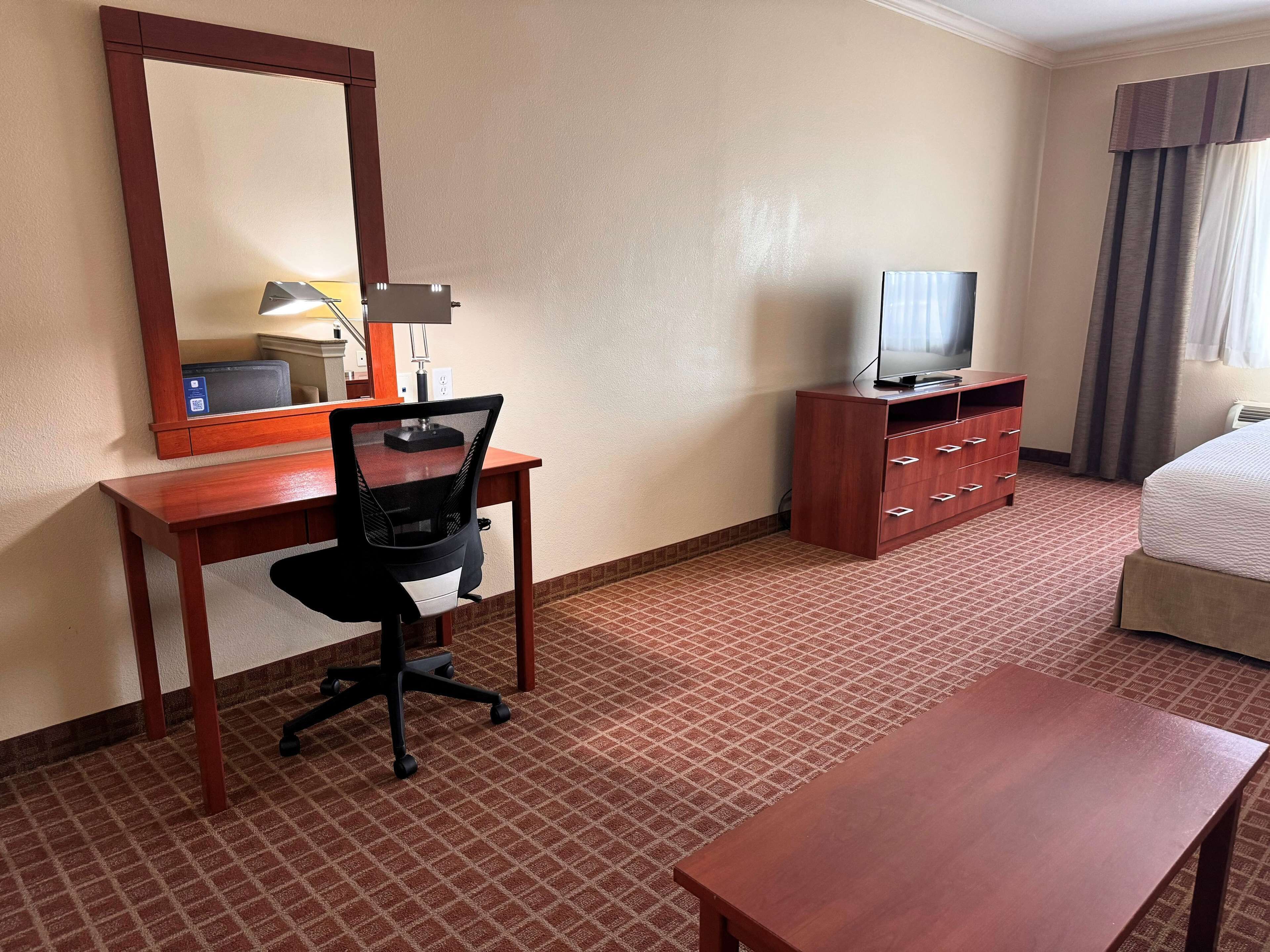 A hotel room with a wooden desk, a black office chair, a television on a wooden stand, a bed with a white comforter, and a large mirror on the wall.