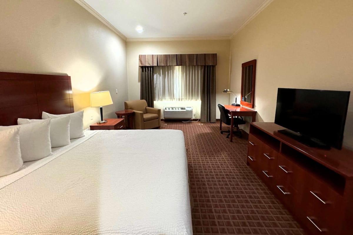 Une chambre d’hôtel avec un grand lit, un bureau avec une télévision, une chaise, une lampe et une fenêtre avec des rideaux.