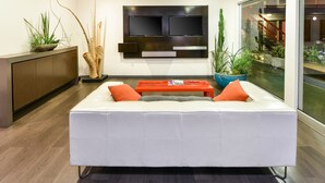 A modern living room with a white sofa, orange cushions, a red coffee table, and a flat-screen TV mounted on the wall. - The Tangerine - A Burbank Hotel (Burbank)