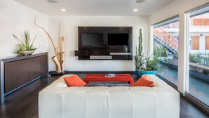 A modern living room with a white sofa, red accents, a flat-screen TV mounted on the wall, and a view of a building with stairs outside. - The Tangerine - A Burbank Hotel (Burbank)