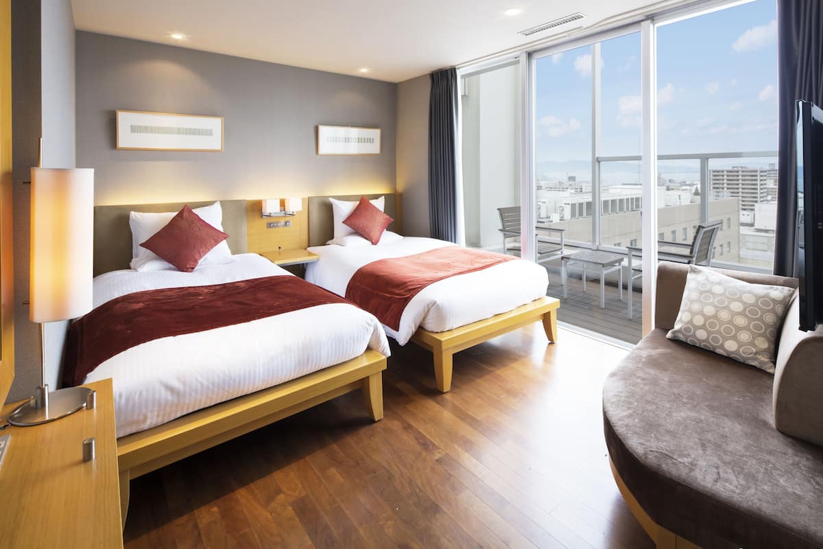 A hotel room with two beds, a sofa, a balcony with a city view, and a large window.