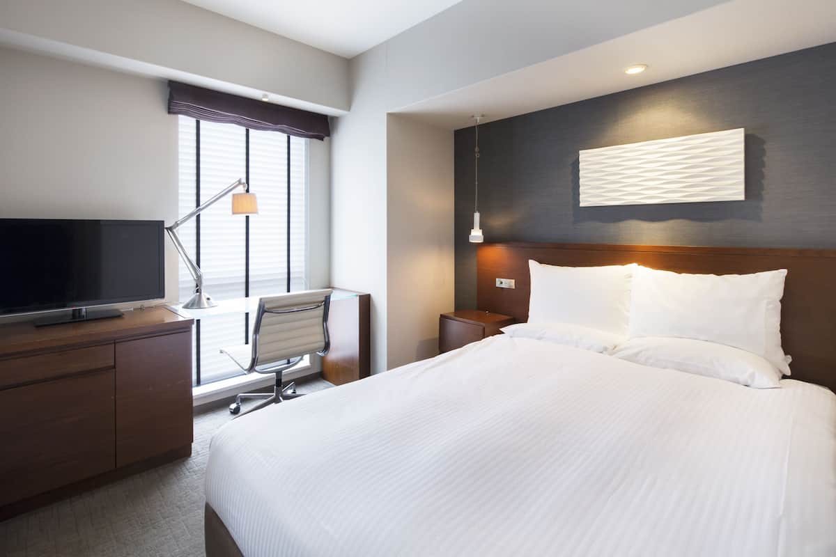 A hotel room with a bed, a desk with a TV, a chair, a nightstand, and a wall-mounted lamp.