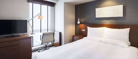 A hotel room with a bed, a desk with a TV, a chair, a nightstand, and a wall-mounted lamp.