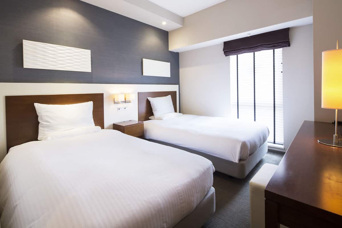 A hotel room with two beds, a wooden headboard, a bedside table with a lamp, and a window with blinds.