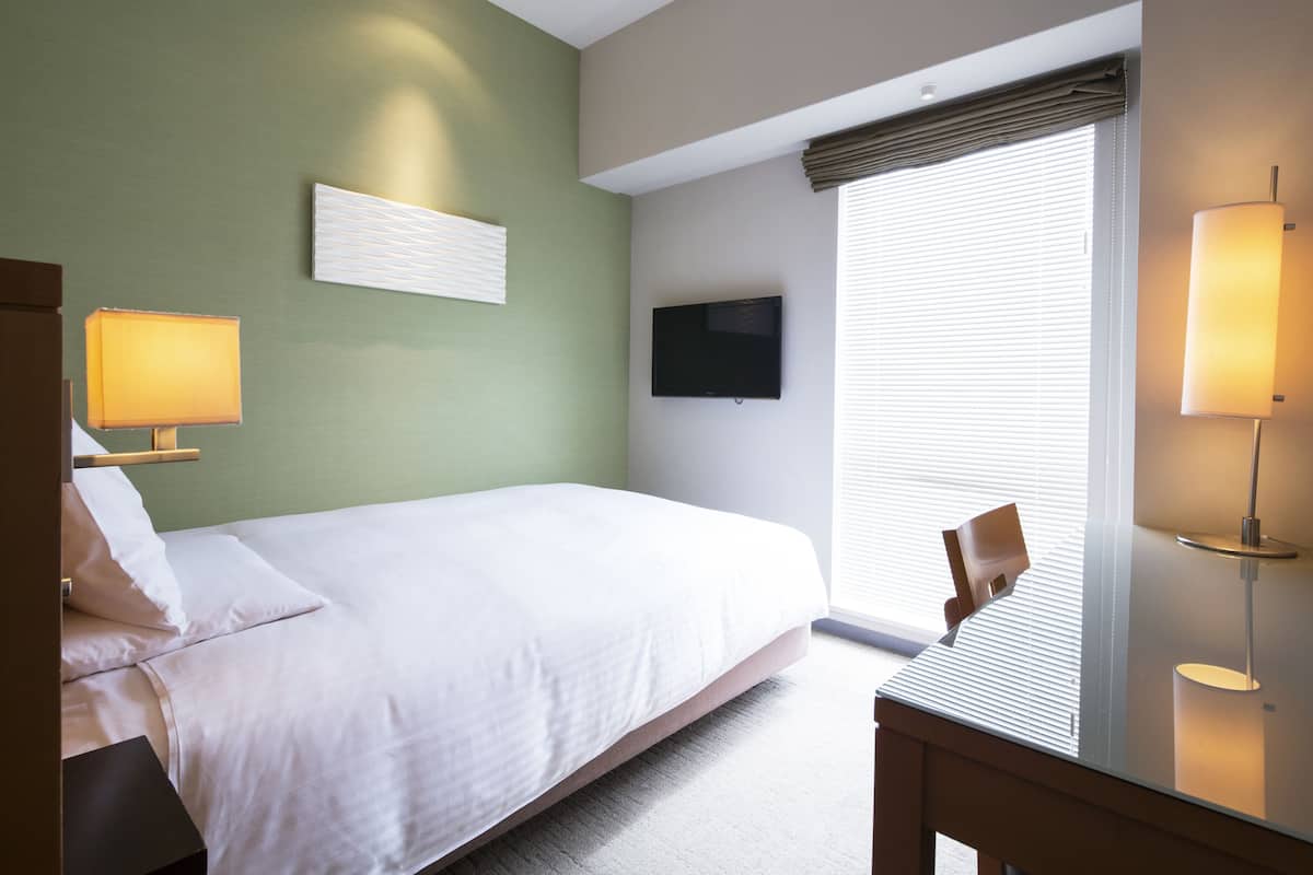 A hotel room with a bed, a desk, a chair, a TV, and a window with blinds.