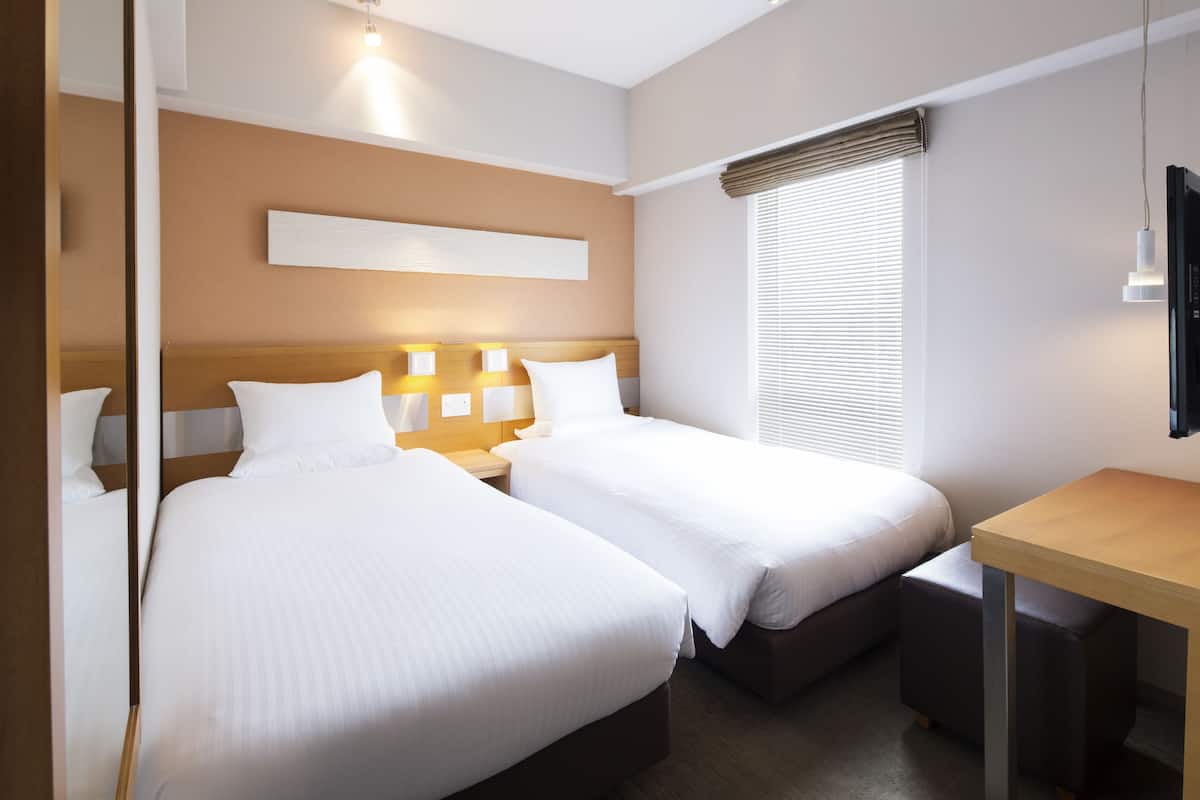 A hotel room with two beds, a wooden nightstand, a small table, and a window with blinds.