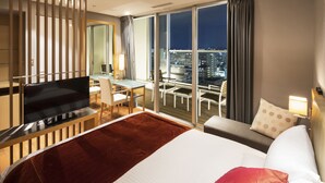 Een hotelkamer met een groot bed, een bureau met een televisie, een balkon met uitzicht op de stad en een modern interieur.