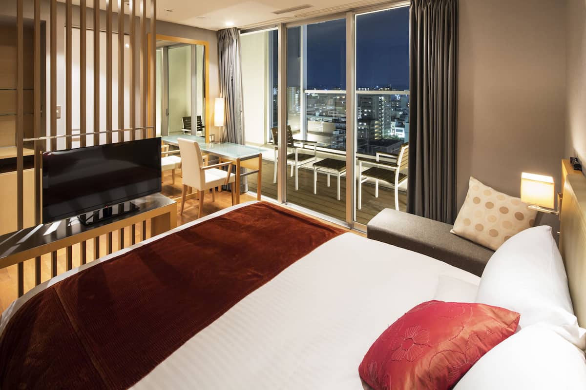A hotel room with a large bed, a desk with a TV, a balcony with a city view, and a modern interior design.