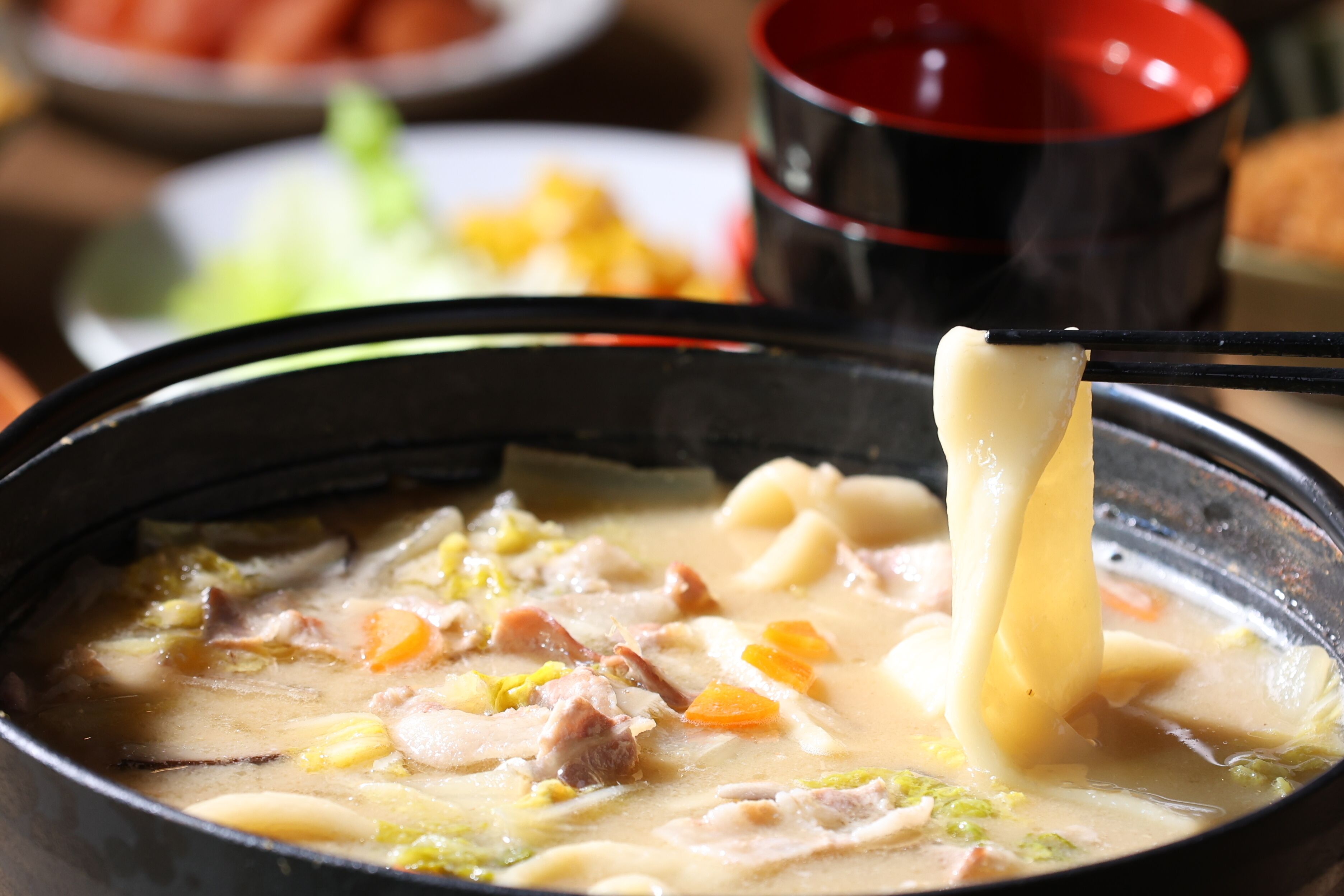 A bowl of soup with various ingredients, including meat and vegetables, being held with chopsticks.