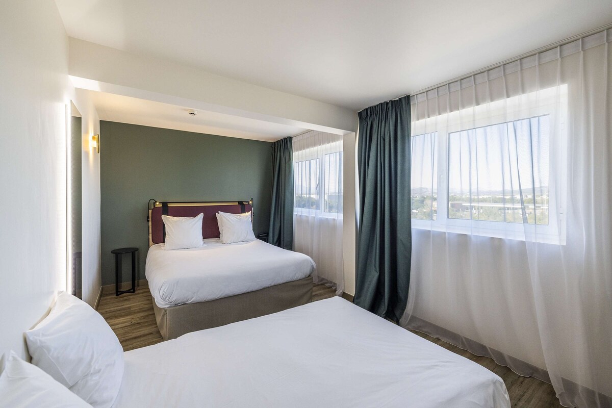 Habitación de hotel con dos camas, vista a la ciudad y una ventana con cortinas traslúcidas.