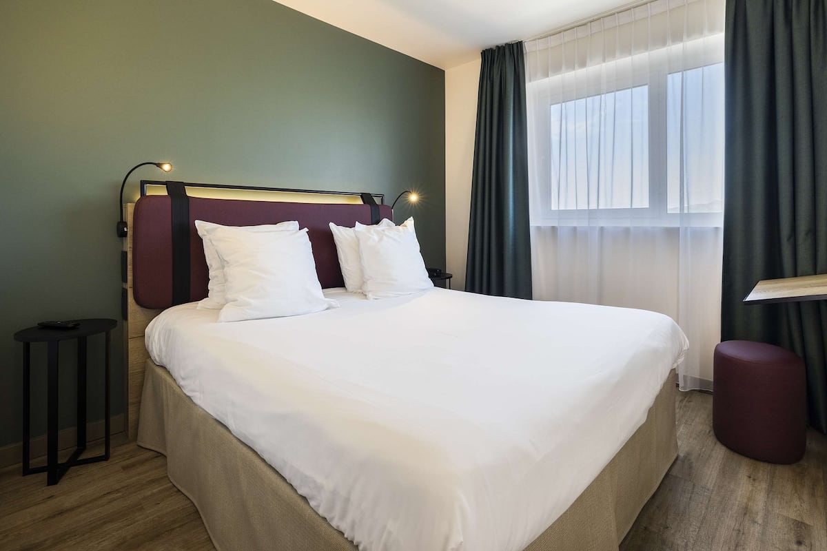 Een netjes opgemaakt bed met witte lakens, een hoofdbord met een lamp en een klein nachtkastje, en een raam met gordijnen.