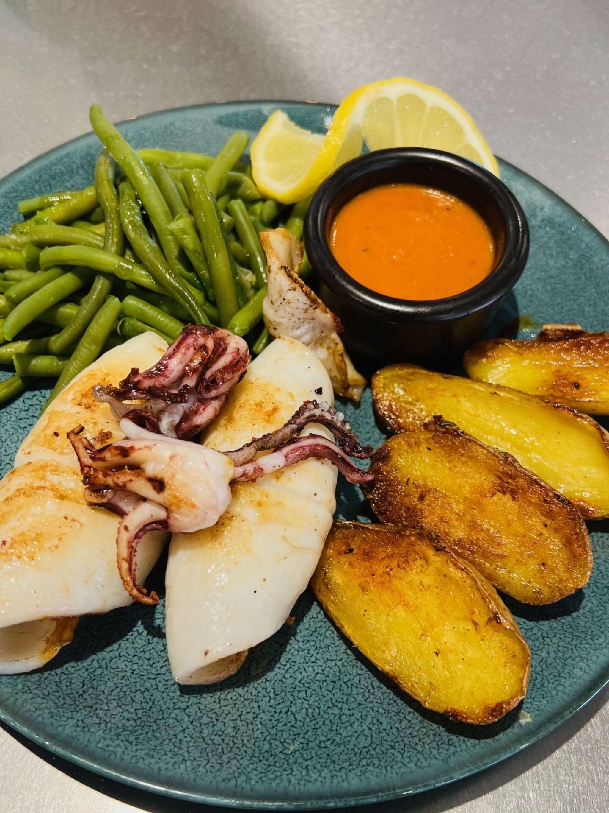 Un plato con pulpo a la plancha, judías verdes, patatas asadas y un gajo de limón, acompañado de una pequeña salsa.